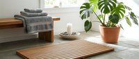 Wooden slat bath mat on gray floor with potted plant, towels, and candles in bright minimalist bathroom interior.