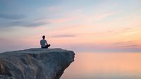 Silhouette of person sitting in meditation pose on cliff edge overlooking calm ocean during peaceful sunset with pink and purple sky. Mindfulness concept.