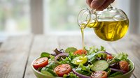 Pouring golden olive oil over fresh salad with tomatoes, mixed greens, and vegetables in white bowl on wooden table for cooking, food preparation, and healthy eating.