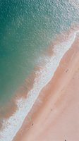 Aerial view of turquoise ocean waves meeting pink sandy beach shoreline, creating dynamic diagonal composition with white foam patterns along waterline.