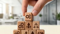Hand holding wooden cube with gear icon on top of pyramid stack of blocks with business symbols in office setting.