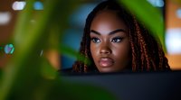 Young African American woman with braided hair and natural makeup gazing through green leaves, creating atmospheric bokeh effect in soft evening lighting.
