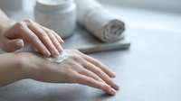 Woman applying moisturizing cream on hands with cosmetic jar and towel on white surface for skincare routine and beauty treatment.