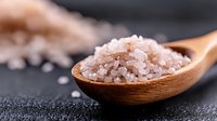 Pink Himalayan salt crystals in wooden spoon on dark background, macro shot with selective focus showing mineral texture and natural gourmet seasoning.