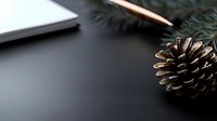 Pine cone and evergreen branch on dark background with notebook and pencil, creating minimalist winter holiday workspace atmosphere for seasonal design projects.