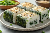 Fresh onigiri sushi rolls with nori seaweed, white rice, and black sesame seeds served on ceramic plate. Green onions and edamame beans as side dishes.