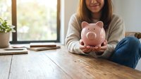 Young Asian woman holding pink piggy bank at wooden table, smiling, representing savings, financial planning, and money management at home.