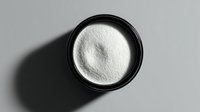 White crystalline powder in black round container on gray background, photographed from above with dramatic shadow for pharmaceutical or cosmetic product presentation.