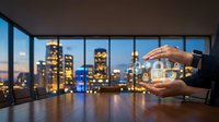 Businessman holding glowing smart home automation device with illuminated cityscape visible through office windows at dusk for technology and real estate concepts.