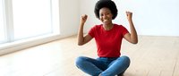 Happy African American woman in red shirt celebrating success sitting cross-legged on wooden floor in bright modern room with raised fists showing joy.