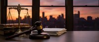 Wooden gavel and scales of justice on desk in law office with city skyline at sunset, representing legal profession and courthouse authority.