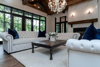 Luxurious living room with white tufted chesterfield sofas, navy velvet pillows, crystal chandelier, exposed wooden beams, and large windows overlooking green landscape.