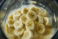 Fresh sliced banana pieces in blender jar with creamy liquid, close-up view showing detailed fruit texture and natural pattern for smoothie preparation.