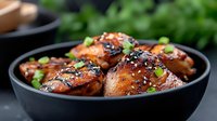 Glazed chicken thighs with sesame seeds and green onions in black bowl, Asian-inspired dish with caramelized sauce for food photography.