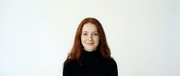 Young redhead woman in black turtleneck sweater smiling at camera against white background. Professional headshot portrait for business marketing.