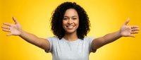 Young woman with curly black hair and medium skin tone wearing gray t-shirt, arms outstretched in welcoming gesture against bright yellow background, expressing joy and positivity.
