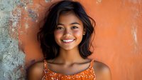 Young Asian woman with natural wavy black hair and bright genuine smile wearing orange floral top against textured orange wall background, radiating joy and confidence.