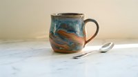 Handcrafted ceramic mug with blue and brown glazed finish on marble surface with silver spoons for coffee tea drinking and kitchen decor.
