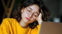 Young Asian woman with glasses in yellow sweater sleeping at laptop, exhausted from work or study in warm evening light.
