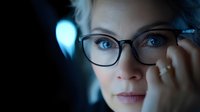 Young Caucasian businesswoman in glasses looking thoughtfully into distance against blue bokeh background, close up portrait showing confidence and intelligence.