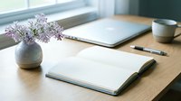Minimalist workspace with open notebook, laptop, coffee cup and purple lilac flowers in ceramic vase on wooden desk near window for productivity and planning.