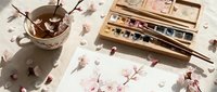 Watercolor painting of cherry blossoms on paper beside a wooden paint palette, brush, and tea cup with pink petals scattered on a linen surface.