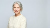 Confident mature woman with gray hair wearing cream cable knit sweater smiling warmly against neutral background for lifestyle and wellness content.