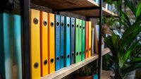 Colorful office binders arranged on wooden shelf with yellow, blue and green folders creating organized rainbow pattern. Indoor workspace with plant decoration.