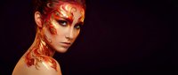 Young Caucasian woman with dramatic phoenix fire body art and face paint in red, orange and gold on dark background.