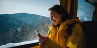 Young woman in yellow jacket using smartphone on train, smiling while traveling through snowy mountain landscape with forest views.