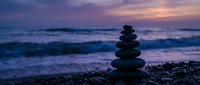 Zen stone stack on pebble beach at sunset with ocean waves and purple sky creating peaceful meditation atmosphere for wellness and mindfulness practices.