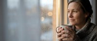 Middle-aged Caucasian woman in cozy sweater enjoying hot beverage by window on rainy day, peaceful morning moment with warm drink and natural light.