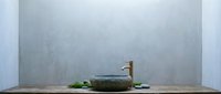 Young woman with East Asian appearance in spa setting, relaxing in natural stone bowl filled with water, surrounded by green leaves and plants against minimalist gray wall background for wellness and relaxation concepts.