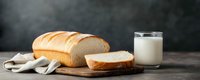 Fresh sliced white bread loaf on wooden cutting board with glass of milk against dark background, rustic food styling for bakery promotion.