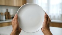Empty white plate held up by male hands with medium skin tone in a bright kitchen setting, suitable for food service, restaurant, culinary, and dining concepts.