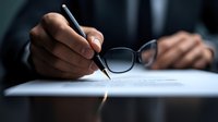 Businessman signing contract document with pen, holding glasses while reviewing legal agreement in corporate office setting.