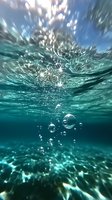 Underwater view of rising air bubbles in turquoise ocean water with sunlight streaming through surface creating dynamic light patterns and reflections.