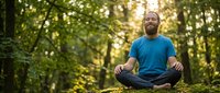 Caucasian male with beard in blue shirt meditating cross-legged in forest clearing surrounded by green trees and natural sunlight, promoting wellness, mindfulness and outdoor relaxation.