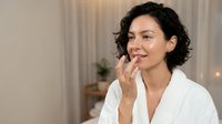 Middle-aged woman with dark curly hair and East Asian ethnicity applying skincare product to face in bright bedroom. Wellness and beauty routine.