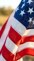 Close-up of waving flag with stars and stripes in sunlight, showing patriotic symbol with red white blue colors and fabric texture details.