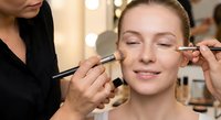Professional makeup artist applying eyeshadow to young Caucasian woman client in modern beauty salon with multiple brushes and cosmetic tools.