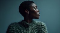 Young Black woman with short natural hair and earrings wearing a textured sage green sweater, looking to the side against a teal background for portrait and fashion use.