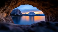 Dramatic mountain lake sunset viewed through natural rock cave opening, with orange sky reflection on calm water surface and silhouetted peaks in distance.