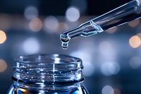 Glass dropper with essential oil or serum drop falling into bottle against blurred bokeh lights background, macro close up wellness concept.