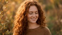 Young redhead woman with curly hair smiling peacefully with closed eyes in warm autumn sunlight wearing cozy brown sweater for lifestyle and wellness content.