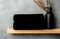 Black rolled towels and matte black bottle with dried flowers on wooden shelf against gray textured wall, minimalist spa bathroom decor composition.