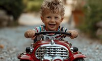 Cheerful curly-haired toddler boy with bright smile riding red toy car outdoors, expressing pure joy and excitement during playtime on summer day.