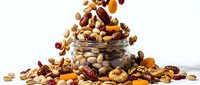 Assorted nuts and dried fruits pouring from glass jar onto white surface, including almonds, cashews, raisins and apricots for healthy snacking.