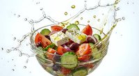 Fresh Greek salad with tomatoes, cucumbers, olives and feta cheese splashing in transparent glass bowl with water droplets on white background.