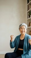Senior Asian woman with short white hair, wearing teal cardigan and black top, smiling with fists raised in confident victory pose against neutral beige wall with shelving, suitable for wellness, motivation, and lifestyle content.